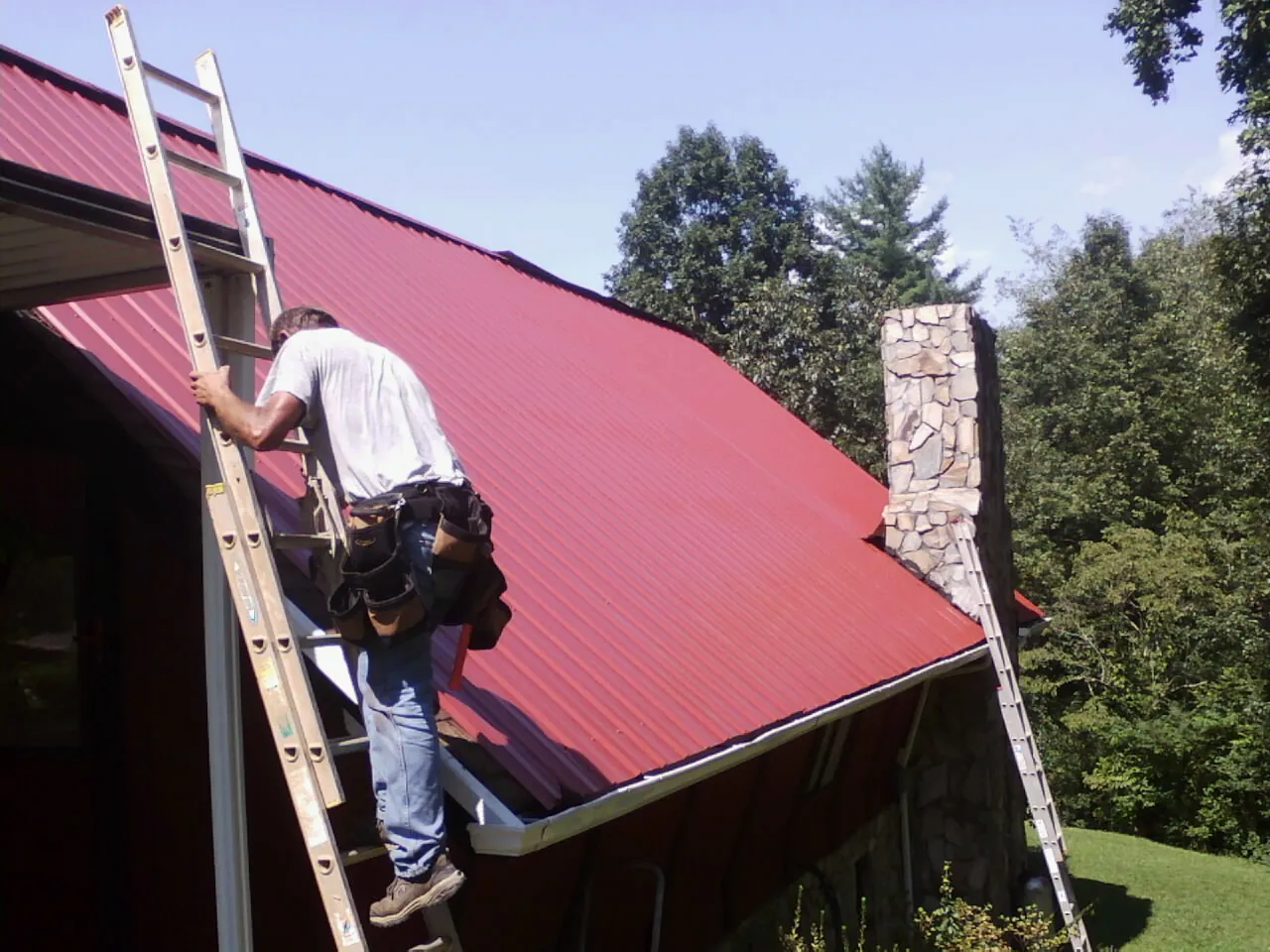 Roofer on a ladder beside a red metal roof and stone chimney — Soffit & Fascia Repair by Roofing Brothers in Wauregan
